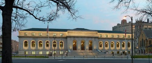 Historic building with large arched windows, illuminated at dusk, surrounded by trees and cityscape.