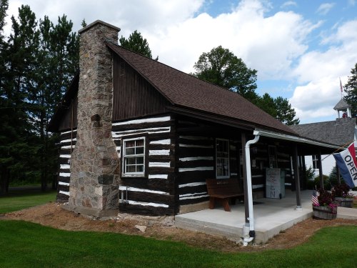 A rustic log cabin with a stone chimney, surrounded by trees and a grassy area, under a partly cloudy sky.