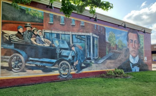 A colorful mural depicting a vintage car with passengers and a man working on it, alongside a portrait of a man in a suit.