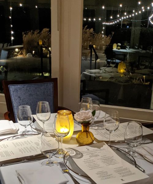 A beautifully set dining table with menus, glasses, and a glowing candle, overlooking a softly lit outdoor scene.