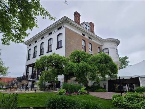 A historic brick building with large windows, surrounded by greenery and a tented area in the garden.