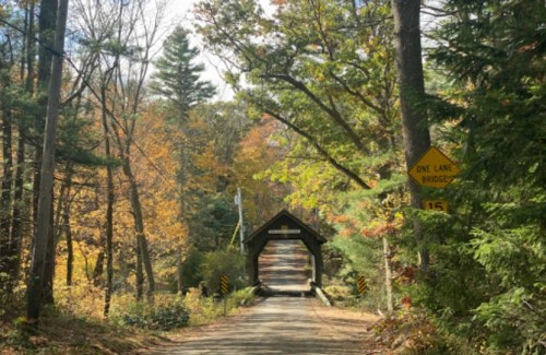 A scenic view of a one-lane covered bridge surrounded by autumn trees and a clear blue sky.