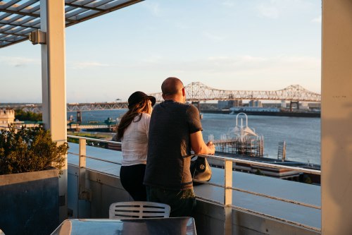 A couple gazes at a river and bridge from a balcony during sunset, enjoying the scenic view.