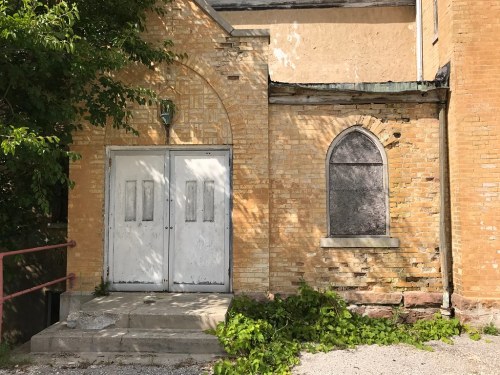Abandoned building entrance with double doors, a boarded window, and overgrown greenery at the base.