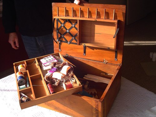 A wooden sewing box with compartments, filled with spools of thread and sewing supplies, partially open.