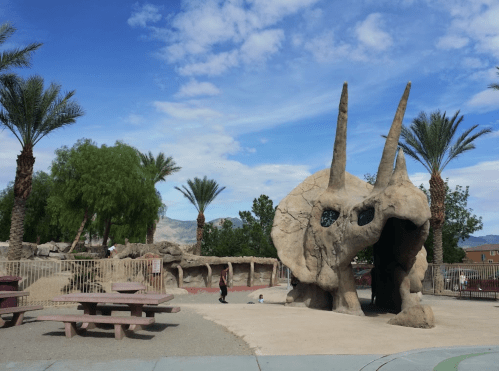 A playground featuring a large dinosaur skull structure, surrounded by palm trees and picnic tables under a blue sky.