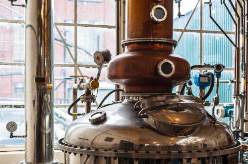 Close-up of a copper and stainless steel distillation apparatus with gauges and pipes, set against a window backdrop.