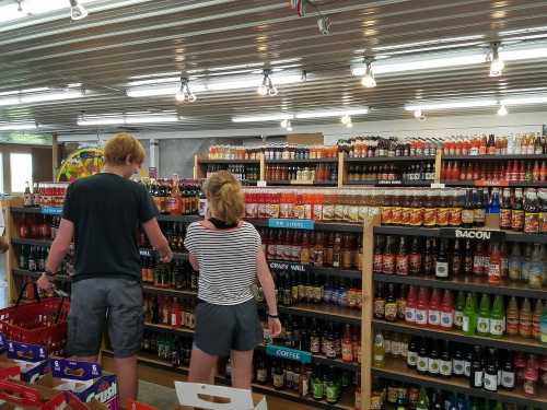 Two people browse a colorful display of various beverages in a store, with shelves filled with bottles in the background.