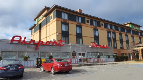 A diner with a red sign reading "Airport Diner" in front of a Holiday Inn hotel, with parked cars in the lot.