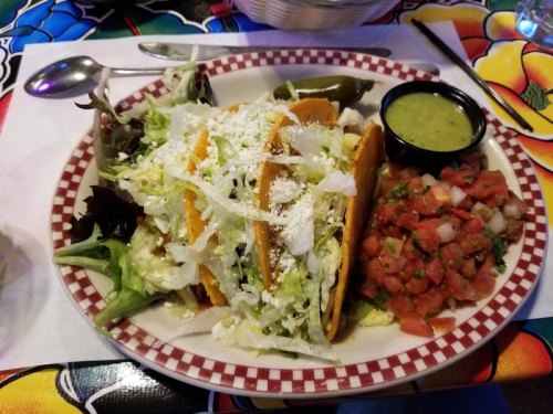 A plate of three tacos topped with lettuce and cheese, served with pico de gallo and a side of green sauce.