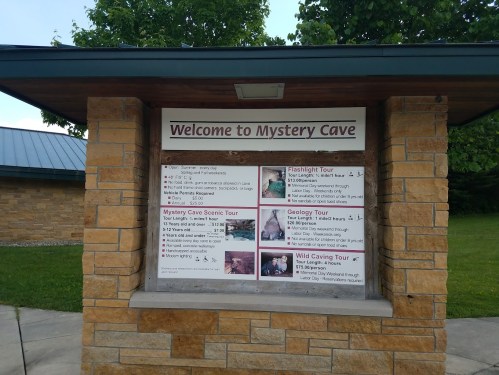 Signboard welcoming visitors to Mystery Cave, listing various tours: Flashlighting, Scenic, Geology, and Wild Caving.