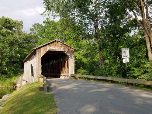 A wooden covered bridge surrounded by greenery, with a weight limit sign nearby and a paved road leading to it.