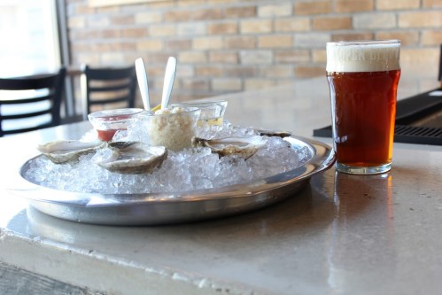 A platter of oysters on ice with sauces, alongside a glass of amber beer on a table.