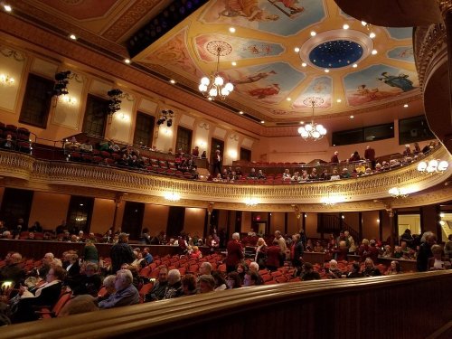Interior of a theater with ornate ceiling, filled with people seated and mingling before a performance.