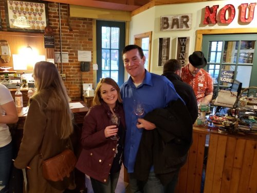 A couple smiles while holding wine glasses at a cozy bar with rustic decor and people in the background.