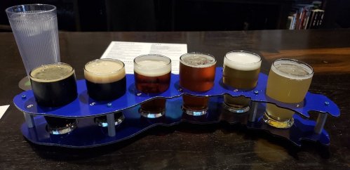 A flight of six craft beers on a blue paddle, with a glass of water beside it, set on a dark wooden table.