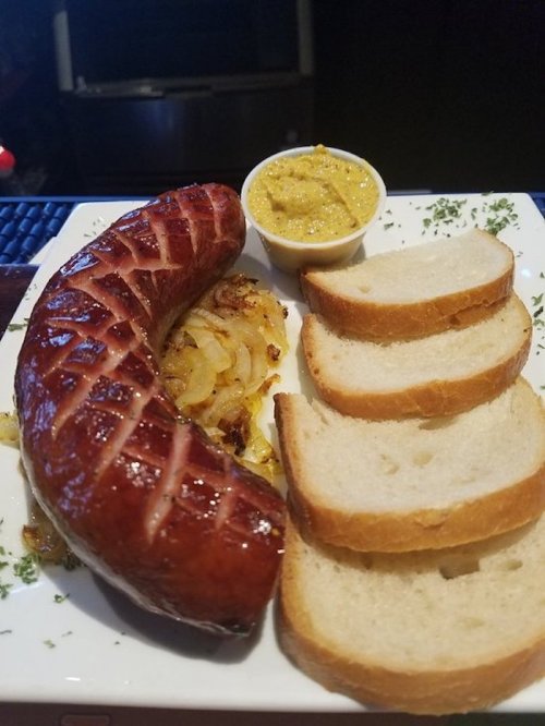A plate featuring a large sausage, sautéed onions, a small cup of mustard, and three slices of bread.