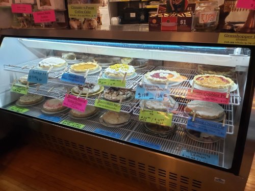 A display case filled with various pies, each labeled with prices and flavors, in a cozy bakery setting.