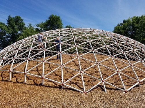 Two children climb on a large, dome-shaped climbing structure made of white pipes, surrounded by trees and blue sky.