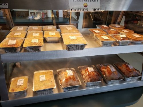A display of various hot food trays in aluminum containers, labeled and arranged on a metal shelf.