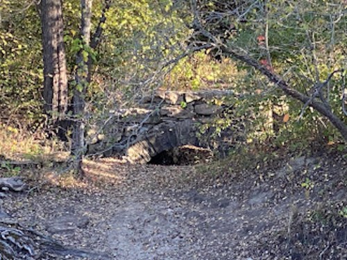 A stone arch bridge surrounded by trees and foliage, with a dirt path leading towards it.