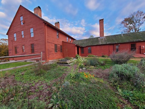 A red wooden house with multiple windows, surrounded by a garden and a blue sky with scattered clouds.