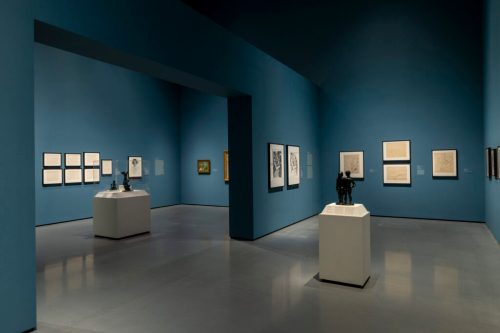 A modern art gallery with blue walls, featuring framed artworks and a sculpture on a pedestal in the center.