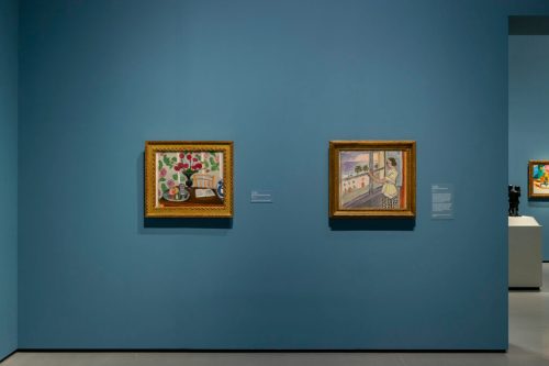 Two framed paintings hang on a blue wall in an art gallery, with labels beneath each artwork.