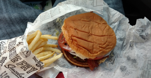 A burger with lettuce, tomato, and bacon next to a serving of French fries, all wrapped in paper.
