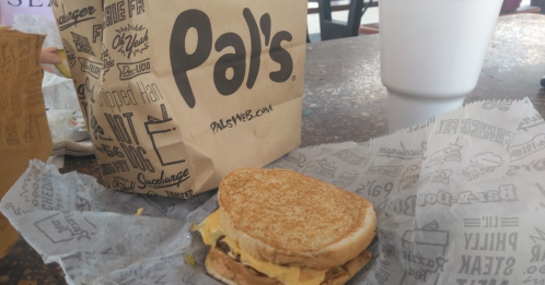 A sandwich on a wrapper next to a takeout bag and a large drink on a table.