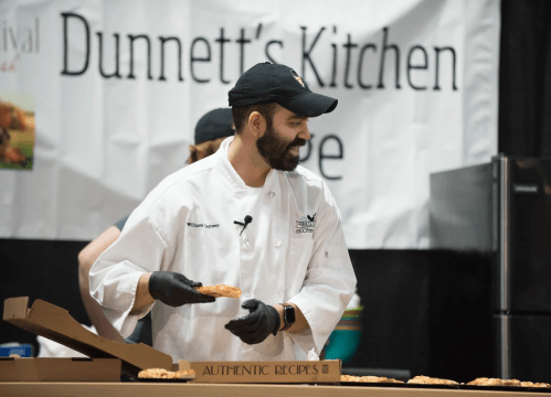 A chef in a white coat and black cap prepares food on stage at a cooking demonstration, smiling and holding a dish.