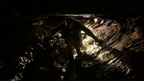 A person wades through a dark, reflective cave, illuminated by a faint light from above.