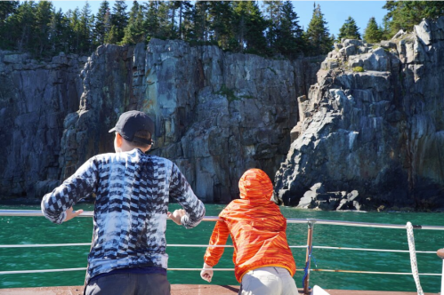 Two people on a boat look out at rocky cliffs and clear blue water on a sunny day.