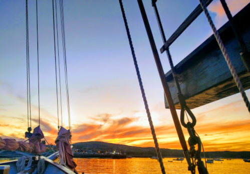 A vibrant sunset over a calm sea, viewed from a boat with ropes and sails in the foreground.