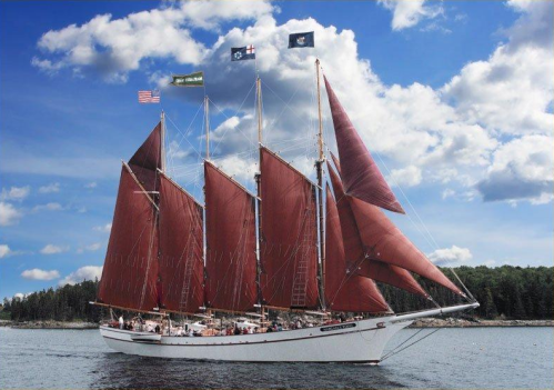 A tall ship with red sails sailing on calm waters under a blue sky with clouds. Flags are flying from the masts.