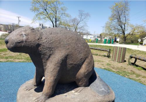 A large stone sculpture of a beaver sits on a grassy area with trees and playground equipment in the background.