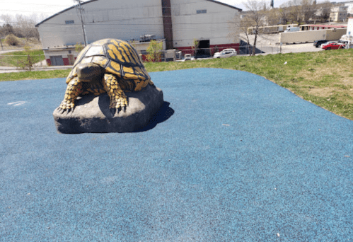 A large turtle sculpture on a blue rubber surface in a park, with a building and vehicles in the background.