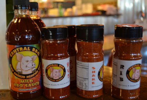 Bottles of Central BBQ mild sauce and various spice rubs lined up on a wooden surface in a restaurant setting.