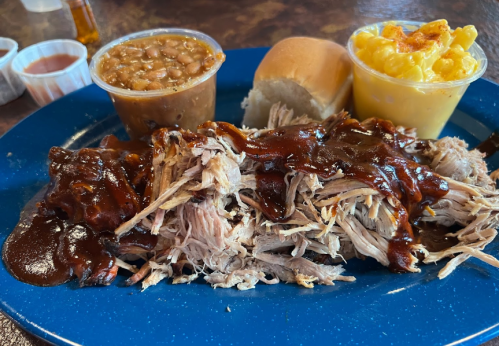 A blue plate with pulled pork in barbecue sauce, baked beans, macaroni and cheese, and a soft roll.