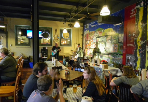 A lively restaurant scene with diners at checkered tables and a band performing in front of a colorful mural.