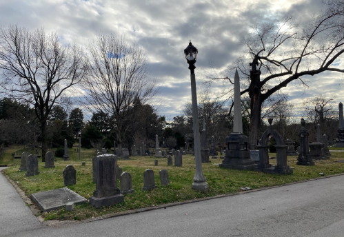 A cemetery scene with gravestones, bare trees, and a lamppost under a cloudy sky.