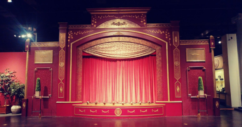 A decorative theater stage with a red curtain, ornate gold details, and potted plants on either side.
