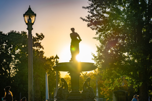 Silhouette of a statue on a fountain at sunset, surrounded by trees and a lamp post.