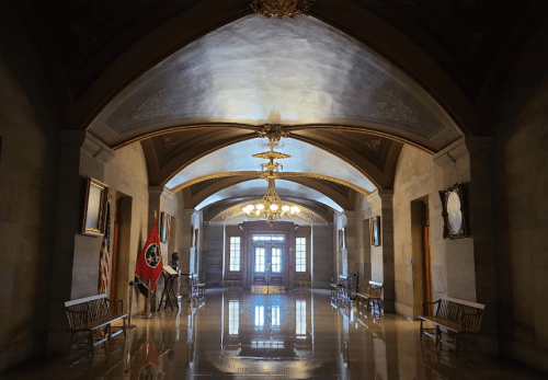 A grand hallway with arched ceilings, flags, and benches, illuminated by a chandelier and natural light.