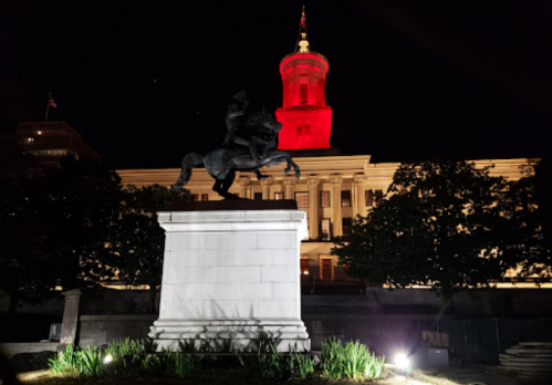 A statue of a horseman in front of a lit building, with the top of the structure illuminated in red at night.