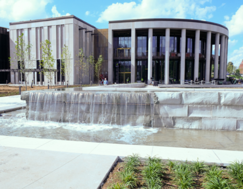 Modern building with large windows beside a waterfall feature and landscaped greenery under a blue sky.