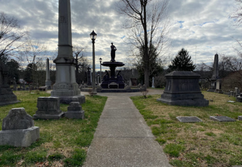 A pathway through a cemetery lined with gravestones and a central fountain under a cloudy sky.