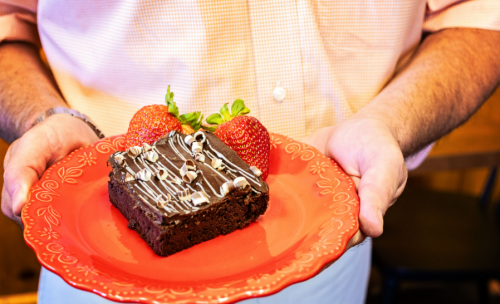 A person holds a red plate with a chocolate brownie topped with drizzled icing and two fresh strawberries.