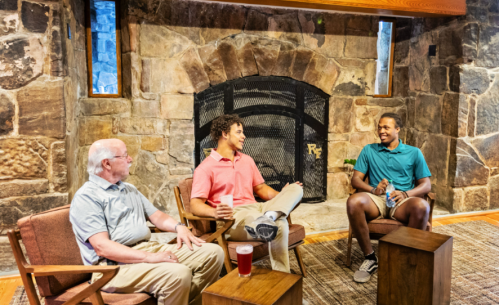 Three men sit in a cozy room with a stone fireplace, enjoying drinks and engaging in conversation.