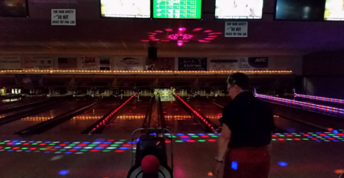 A bowler prepares to roll a ball down a brightly lit bowling alley with colorful lights and screens in the background.